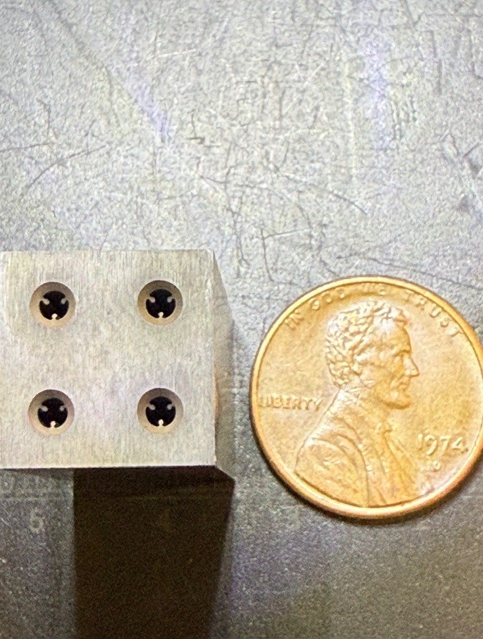 A small precision-machined metal block with four holes, shown next to a penny for scale